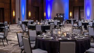 Un salón de banquetes con mesas redondas dispuestas para el servicio de comidas, equipado con sillas, platos, cubiertos y velas.