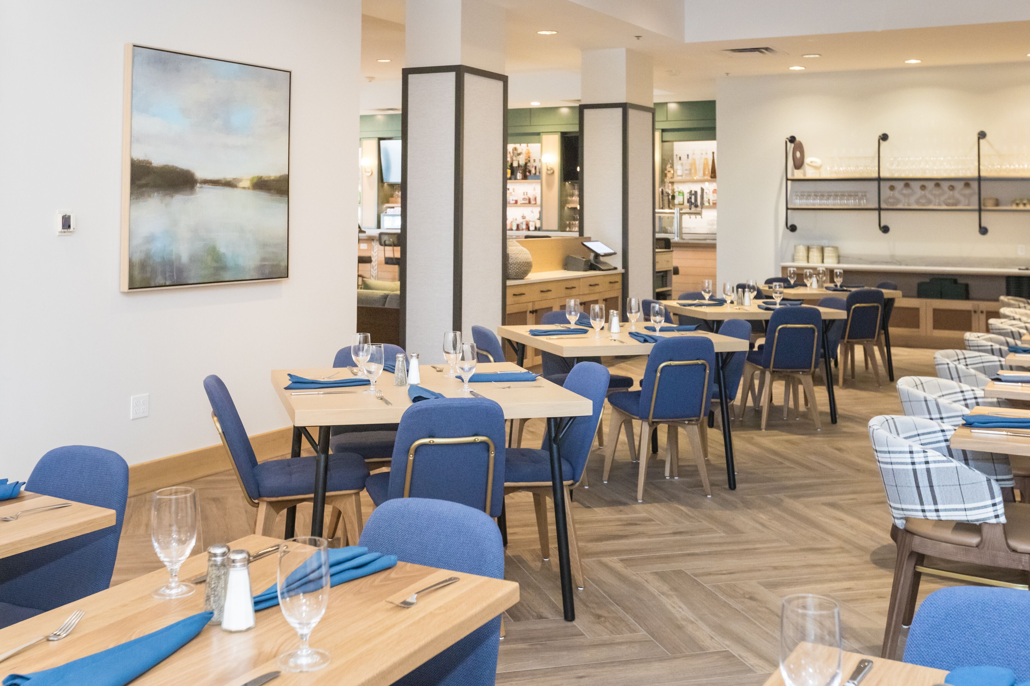 A restaurant with multiple tables, blue chairs, and a bar area in the background.