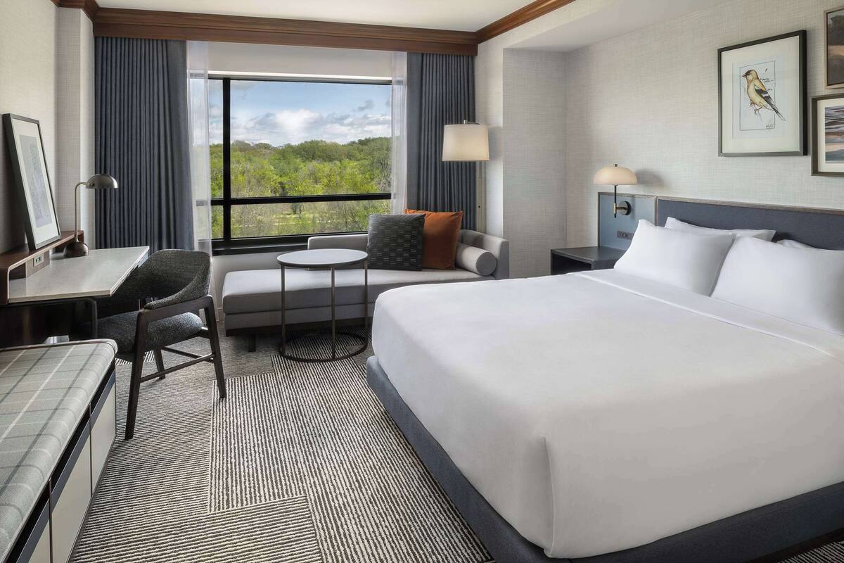Une chambre d’hôtel avec un grand lit, un bureau avec une chaise, une petite table et une fenêtre donnant sur un paysage verdoyant.