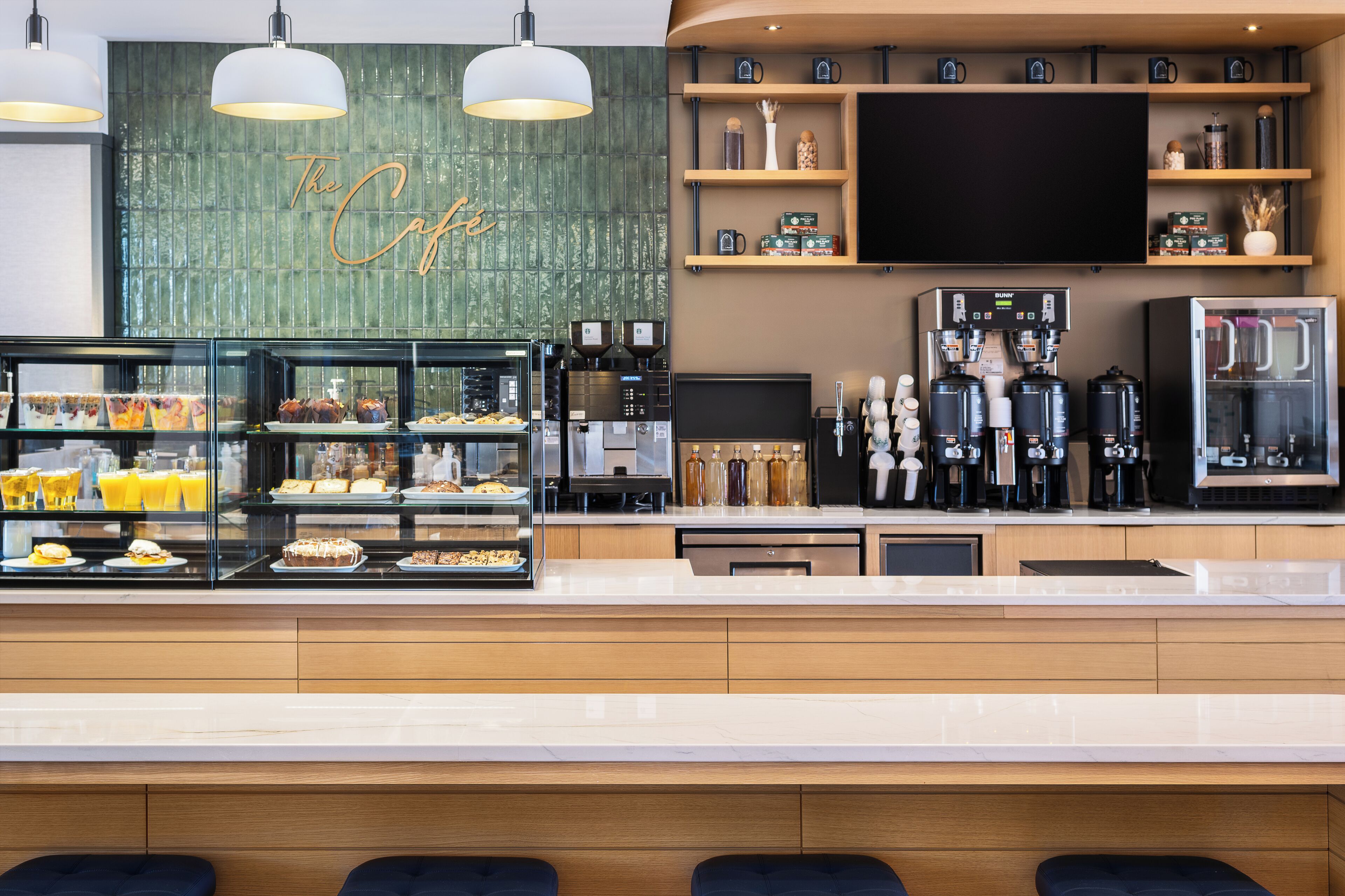 A modern cafe with a glass display case of pastries, a coffee machine, and a flat-screen TV on the wall.