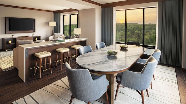 A modern hotel room with a large dining table, comfortable seating, a TV, and a view of the outdoors.