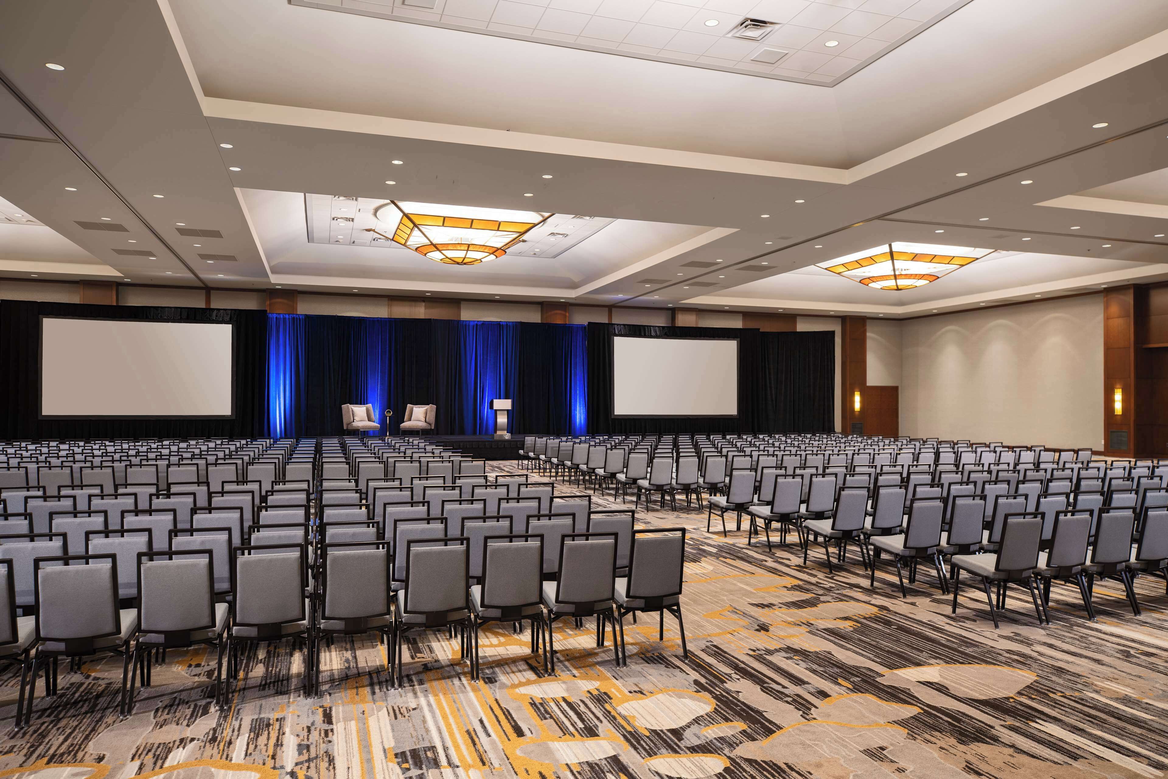 A large conference room with rows of chairs, a stage with a blue curtain, and a large screen.