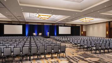 A large conference room with rows of chairs, a stage with a blue curtain, and a large screen.