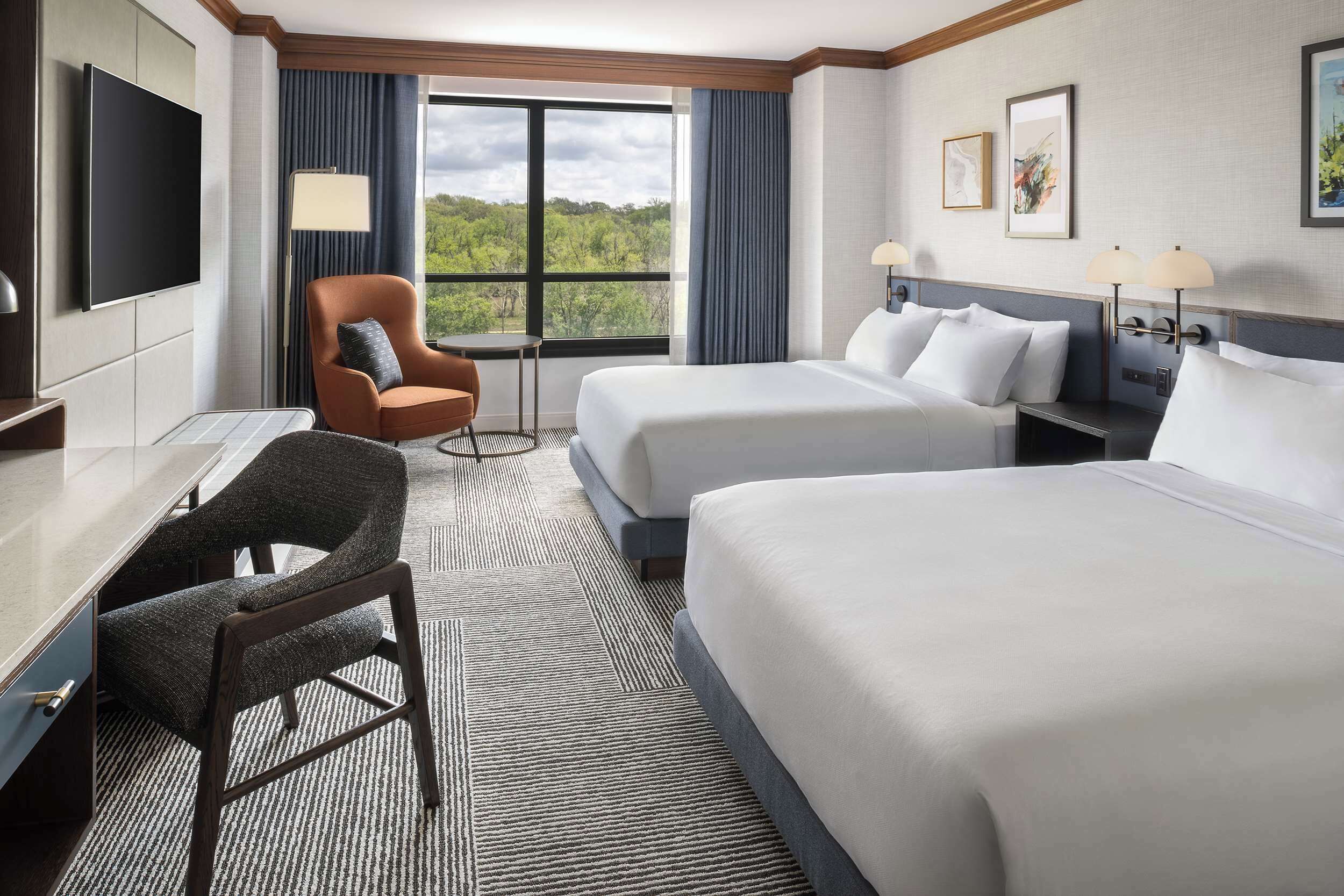 A hotel room with two beds, a desk, a chair, a TV, and a large window with a view of trees.