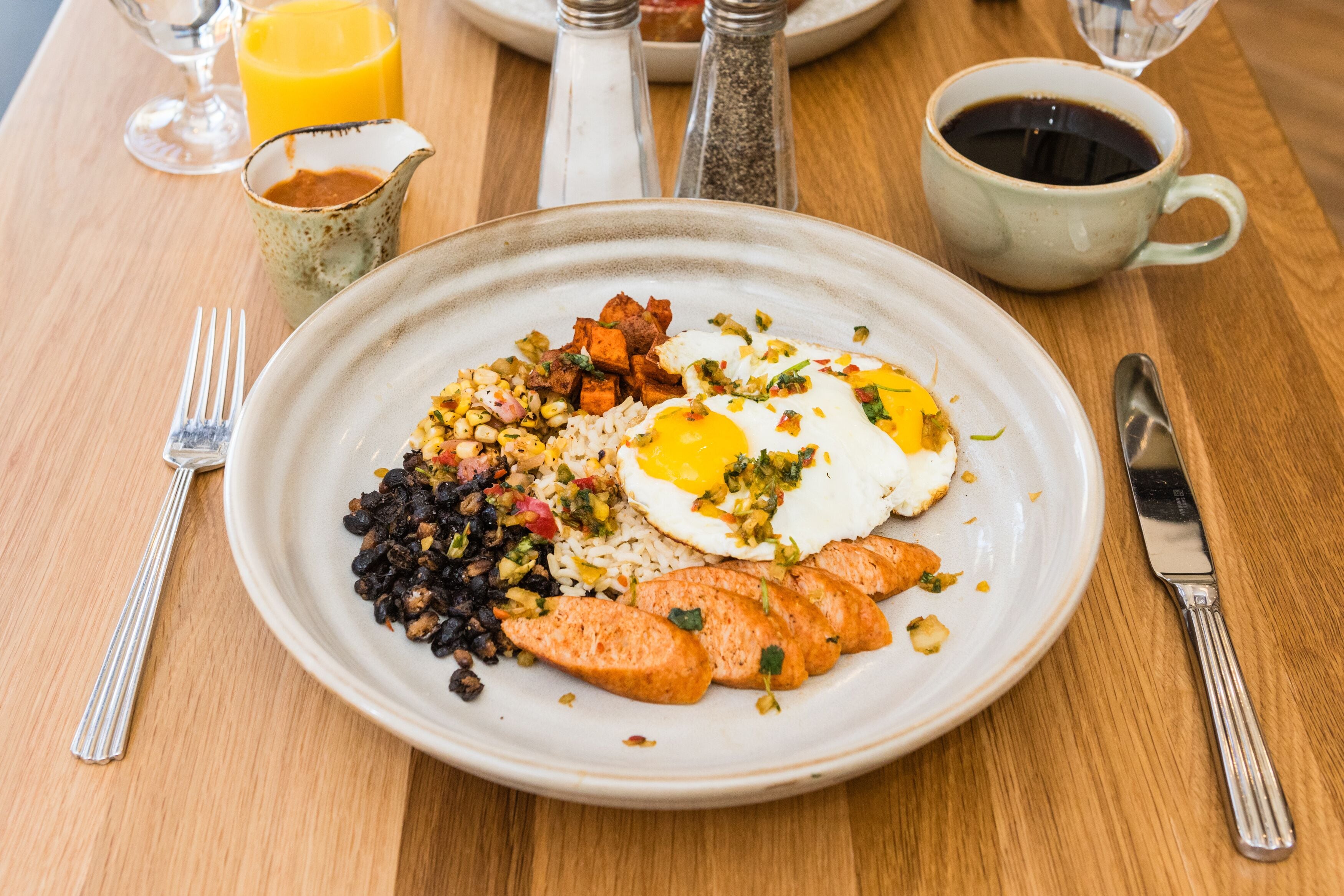 Ein Teller mit Frühstücksleckereien: Eiern, schwarzen Bohnen, Reis und einem Stück Fleisch, dazu eine Tasse Kaffee und ein Glas Orangensaft auf einem Holztisch.