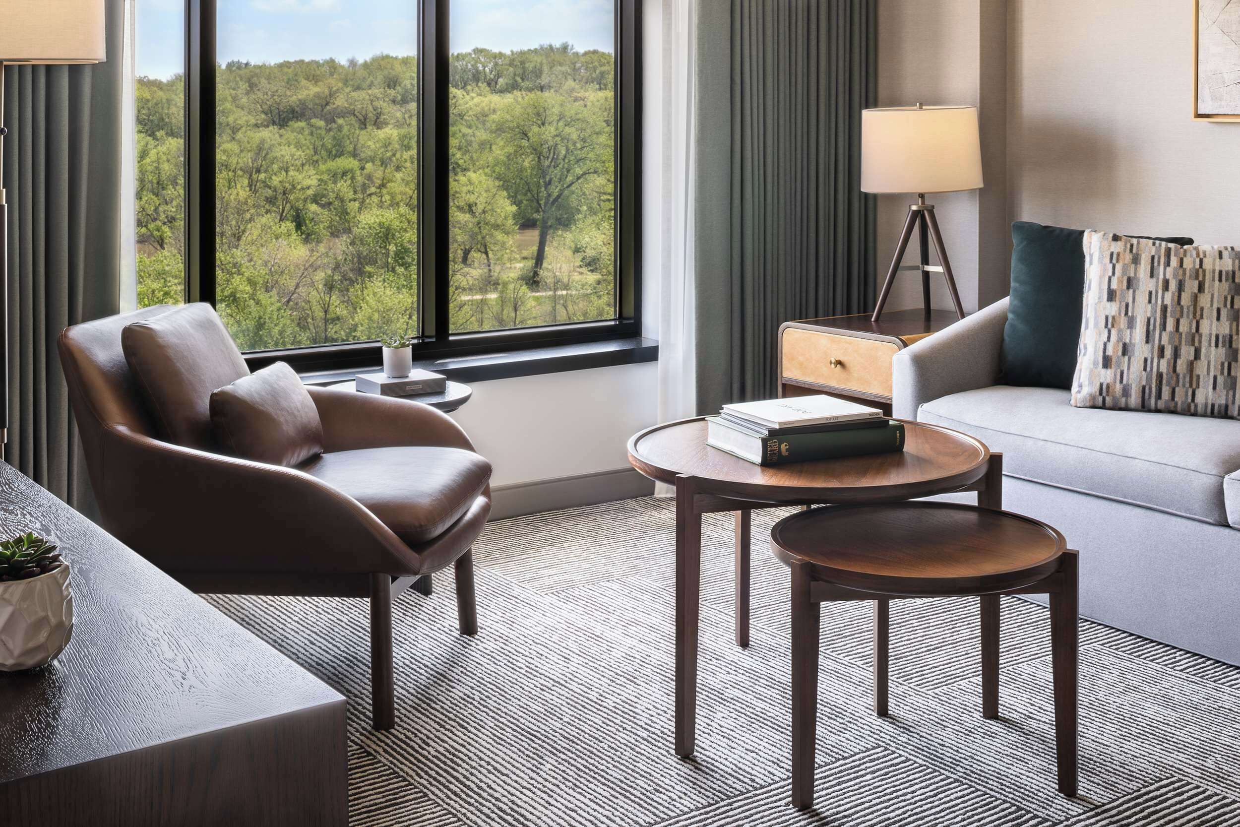 A modern hotel room with a leather armchair, a round wooden table, a sofa, and a view of a forest through large windows.