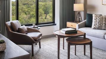 A modern hotel room with a leather armchair, a round wooden table, a sofa, and a view of a forest through large windows.