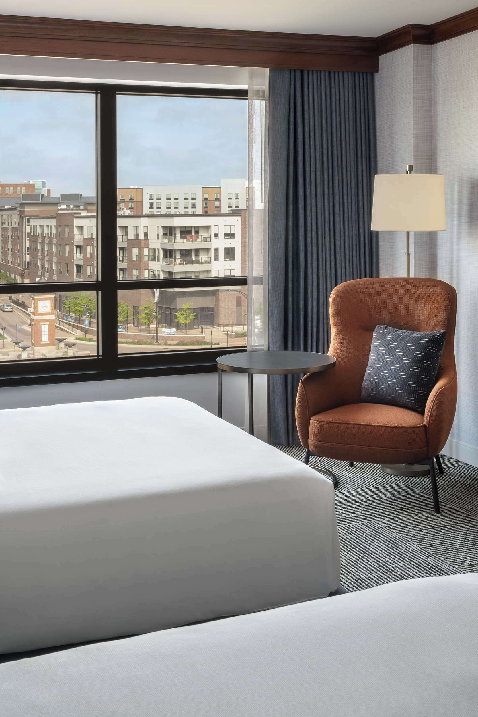 A hotel room with a large bed, a chair with a patterned pillow, a small table, and a view of the city through a large window.