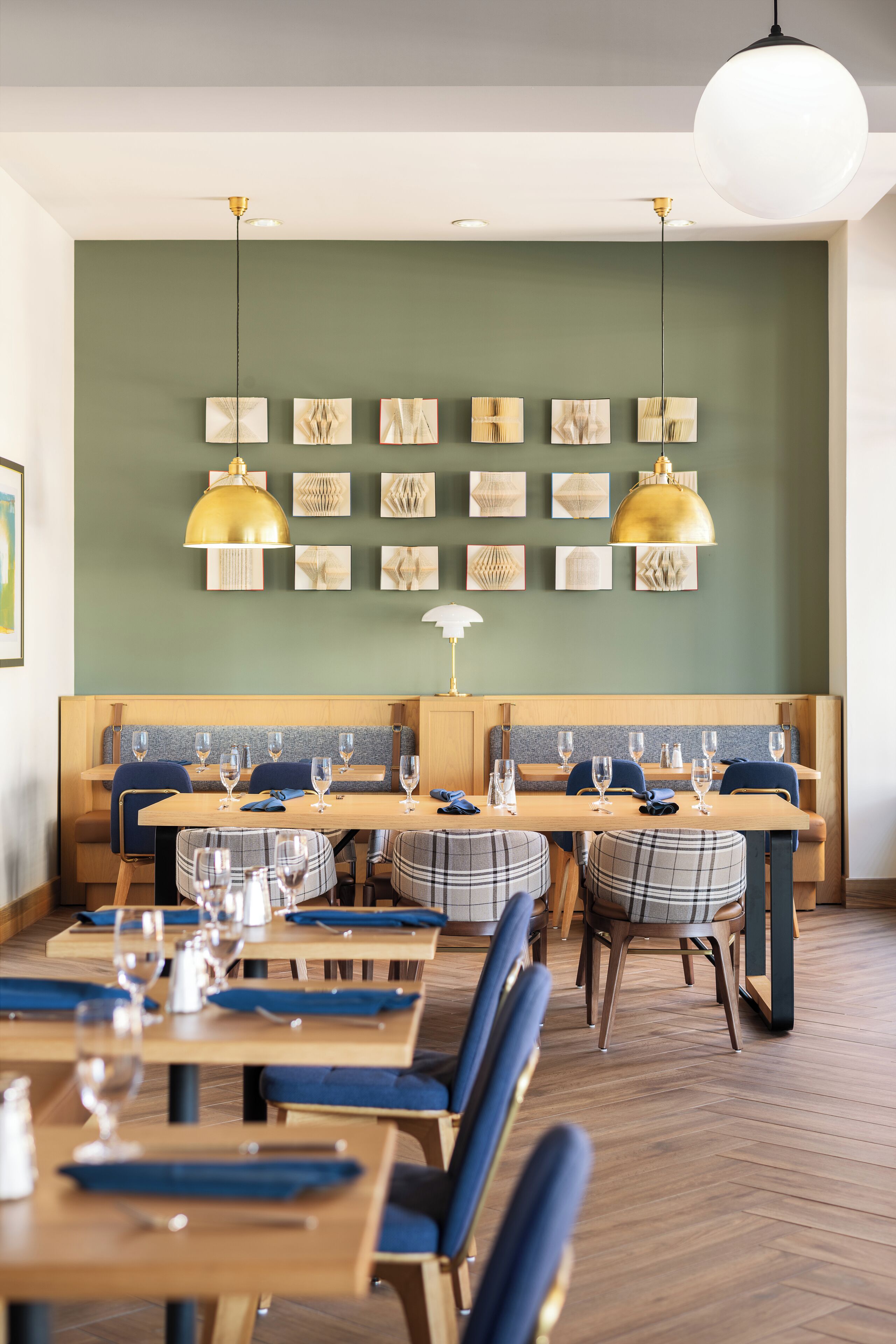 A restaurant with wooden tables and blue upholstered chairs, a green wall with framed artwork, and pendant lighting.