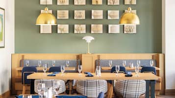 A restaurant with wooden tables and blue upholstered chairs, a green wall with framed artwork, and pendant lighting.