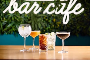 Vier verschiedene Cocktails auf einem Holztisch, im Hintergrund ein Schild mit der Aufschrift 'bar cafe'.
