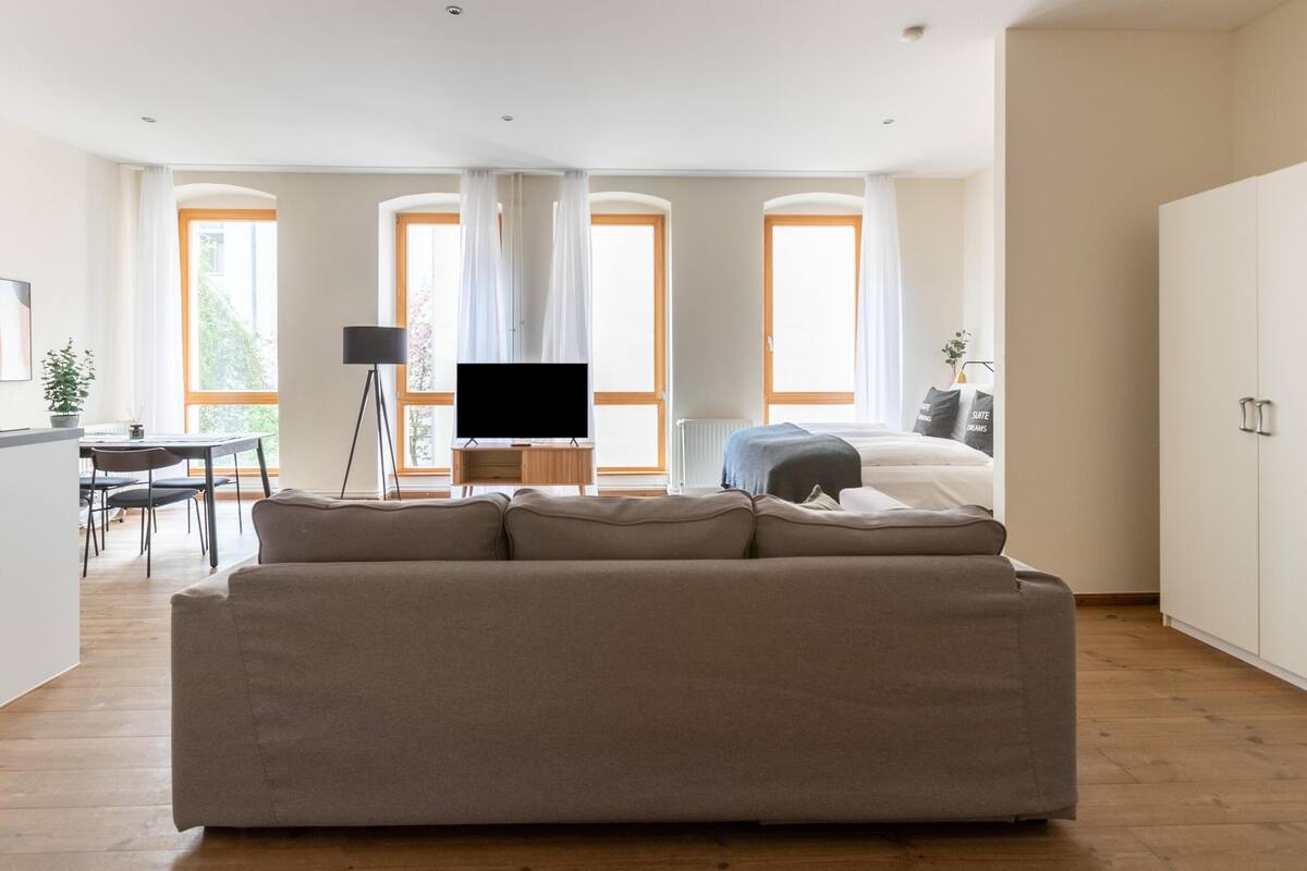 Una sala de estar moderna con un sofá grande, un televisor sobre un mueble bajo y una zona de comedor con una mesa y sillas.