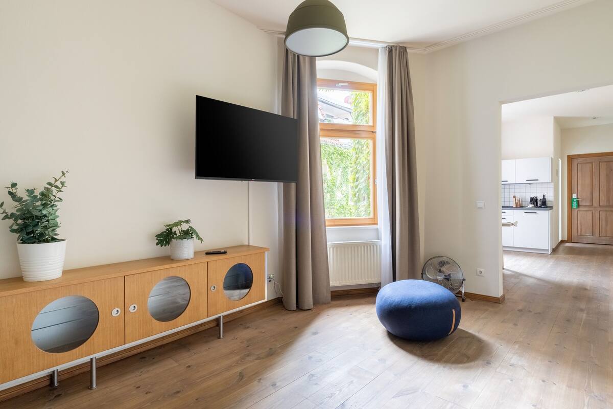 Una sala moderna con un mueble de madera, un televisor, una planta en maceta, un reposapiés azul y vistas a la cocina a través de una ventana.