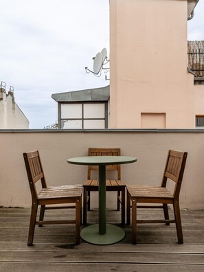 Eine Holzterrasse mit einem runden Tisch und zwei Stühlen, eine beigefarbene Wand und eine Satellitenschüssel im Hintergrund.