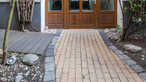 Una puerta doble de madera con paneles de vidrio, un camino de ladrillo y un pequeño jardín con rocas y plantas.
