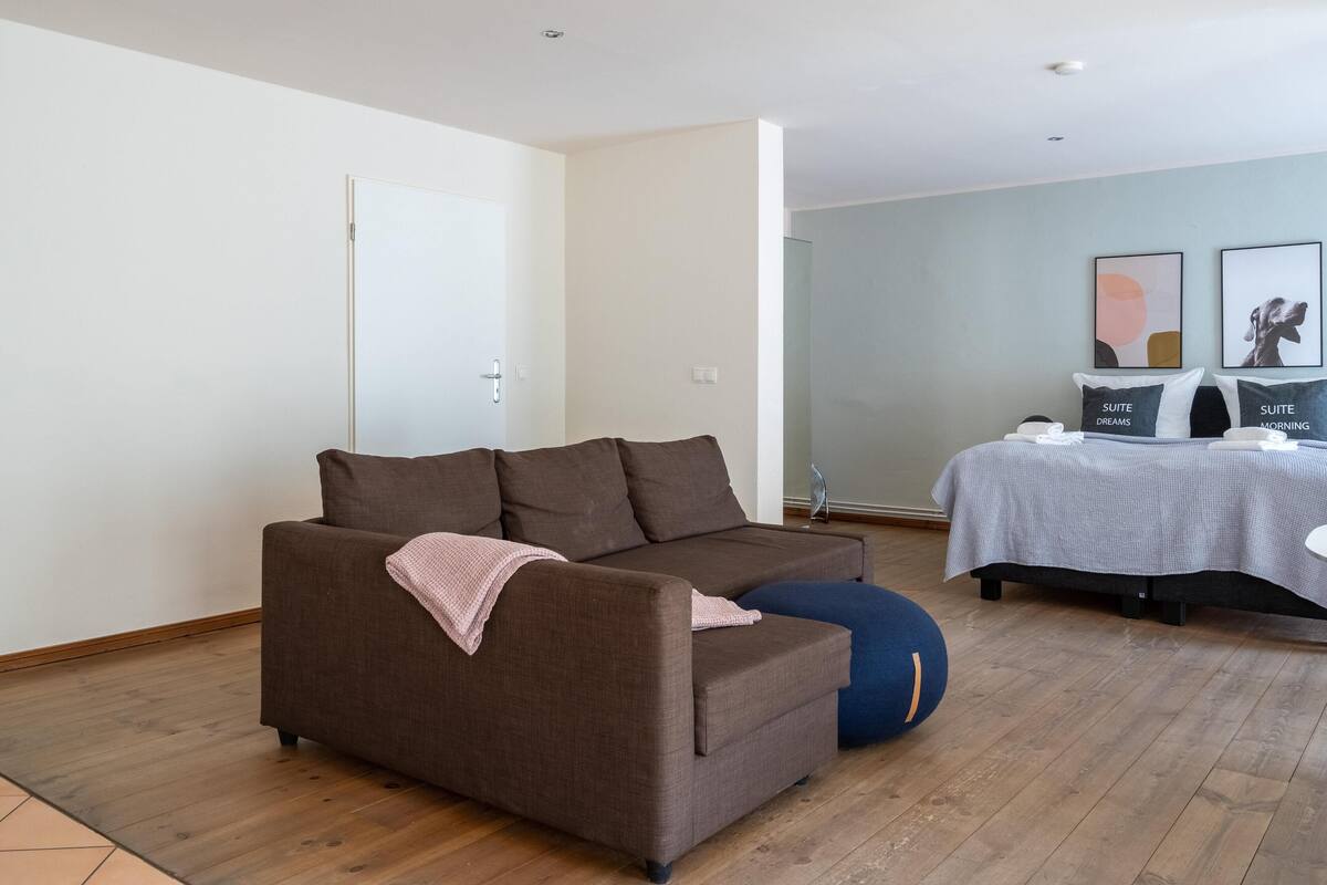 Una sala de estar moderna con un sofá marrón, un reposapiés azul, una cama con cubrecama blanco y dos cuadros en la pared.