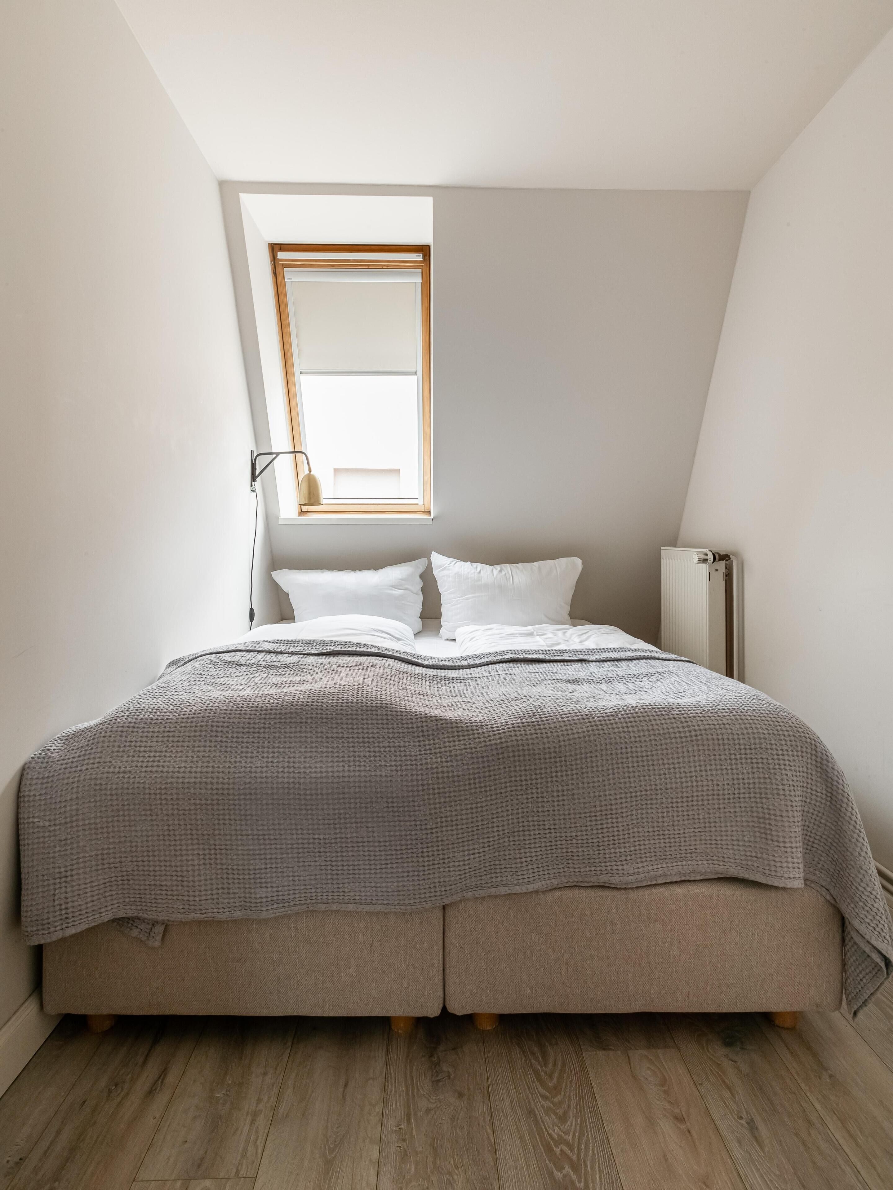 Une chambre avec un plafond en pente, une seule fenêtre, un lit recouvert d’une couverture grise, une lampe de chevet et un radiateur.