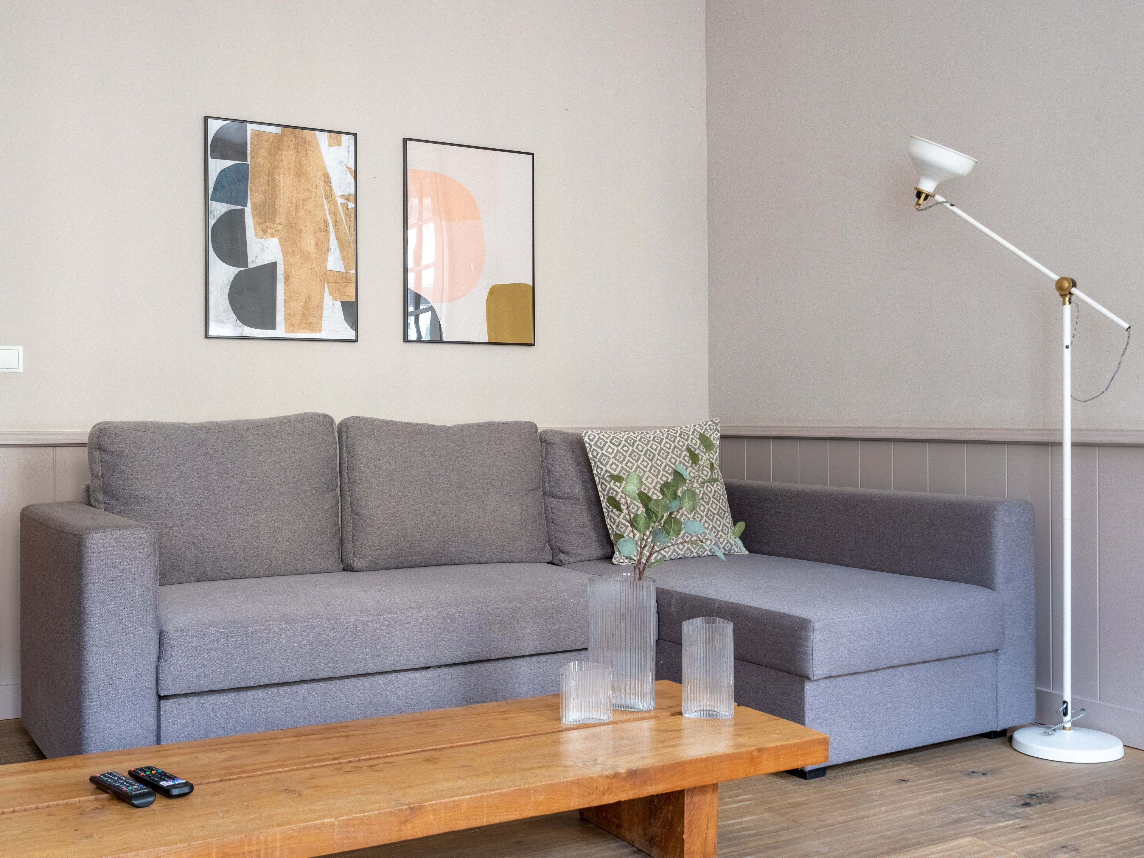 A living room with a grey sofa, two framed abstract artworks, a patterned throw pillow, a wooden coffee table with three clear vases, and a white floor lamp.