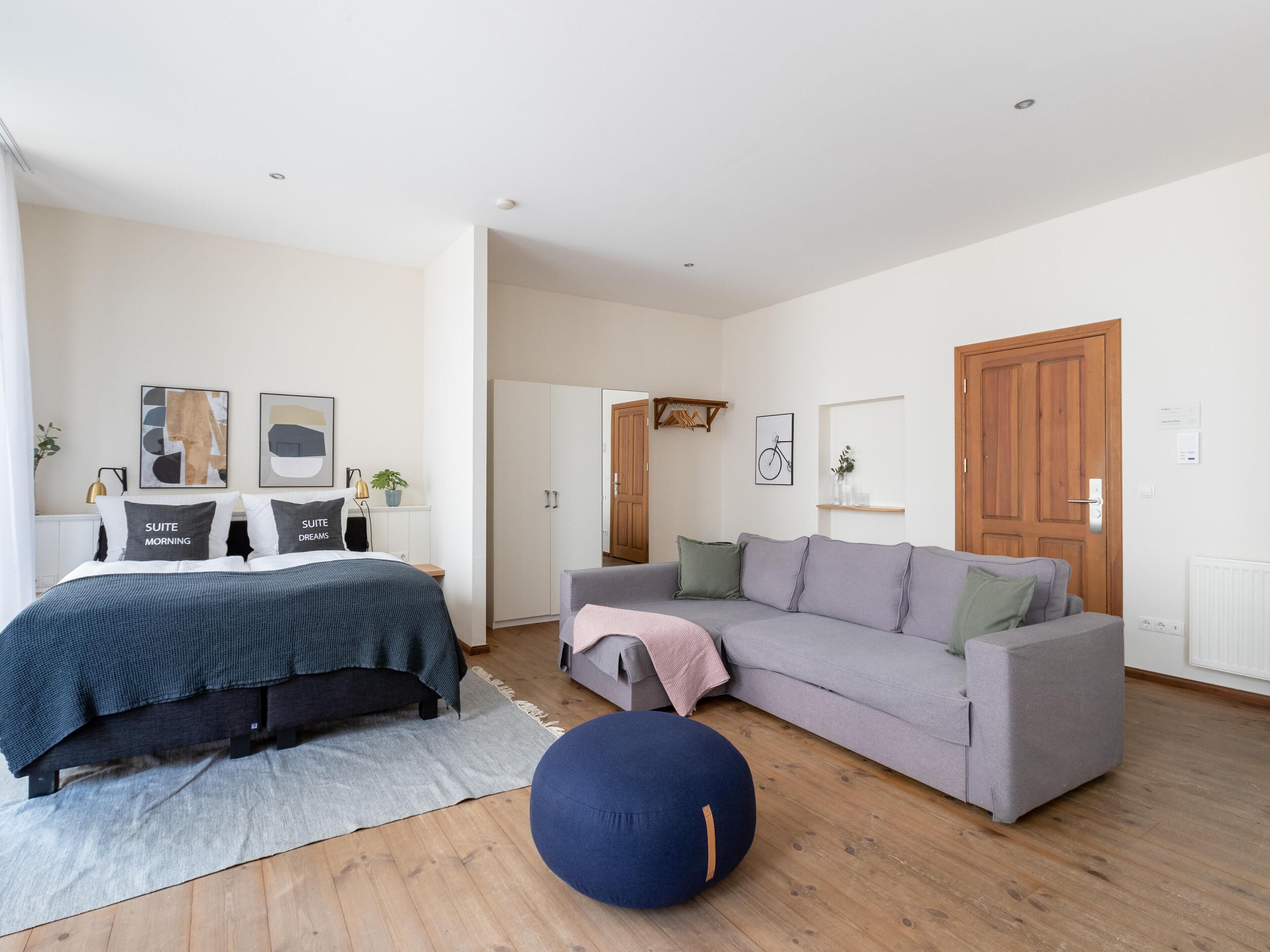 Une chambre moderne avec un lit, un canapé gris, un pouf rond et une armoire.