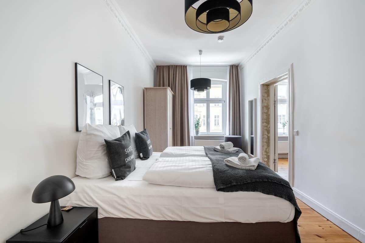 Una habitación de hotel moderna con una cama grande, una mesita de noche con lámpara, un armario y vistas a la ciudad desde la ventana.