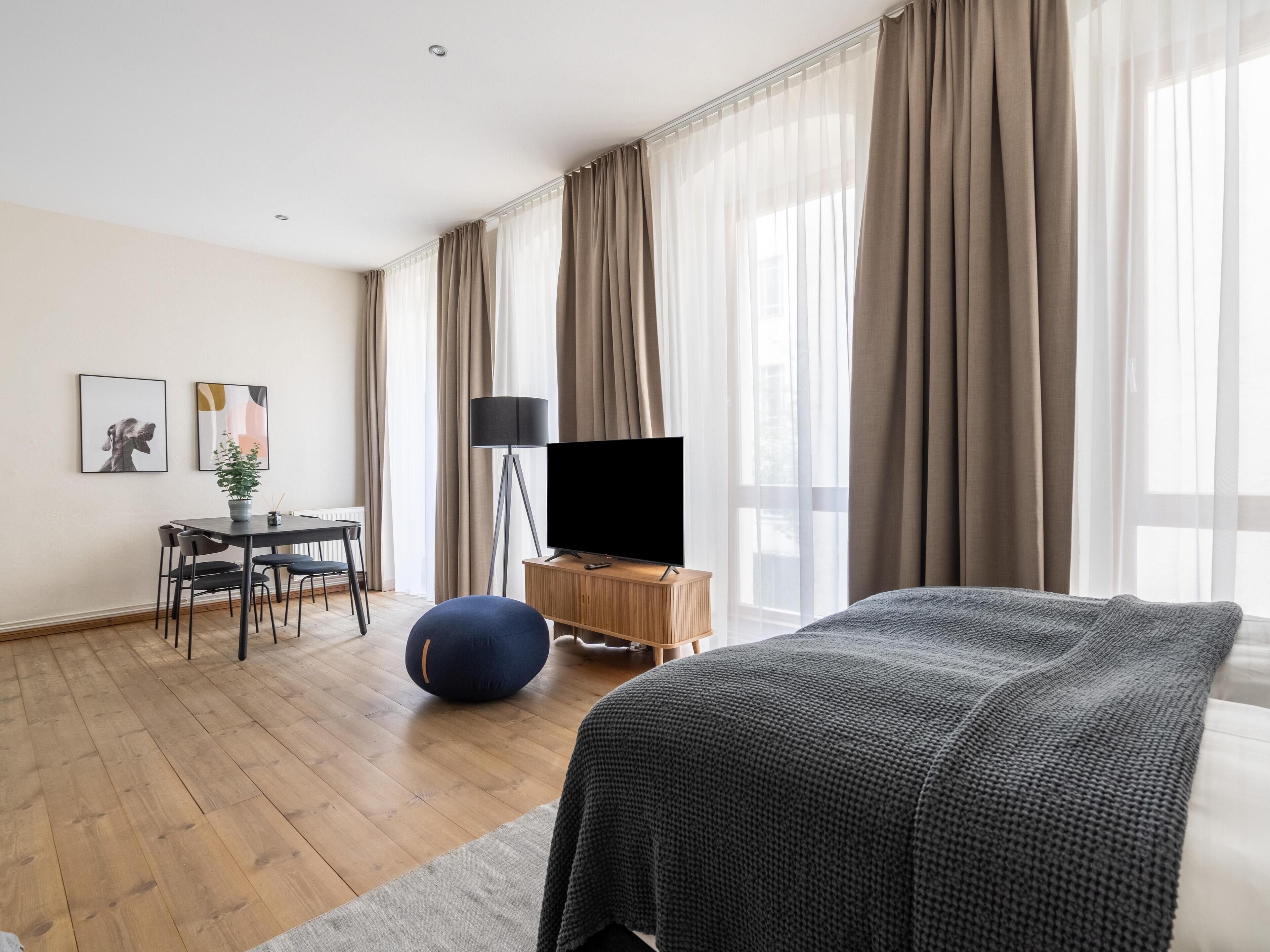 Moderni hotellihuone, jossa on suuri sänky, taulutelevisio, ruokapöytä tuoleineen ja ruukkukasvi.