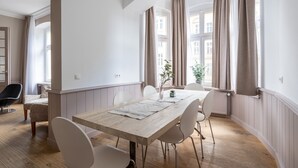 Un comedor con mesa de madera, sillas blancas y vistas a edificios a través de ventanas grandes con cortinas.