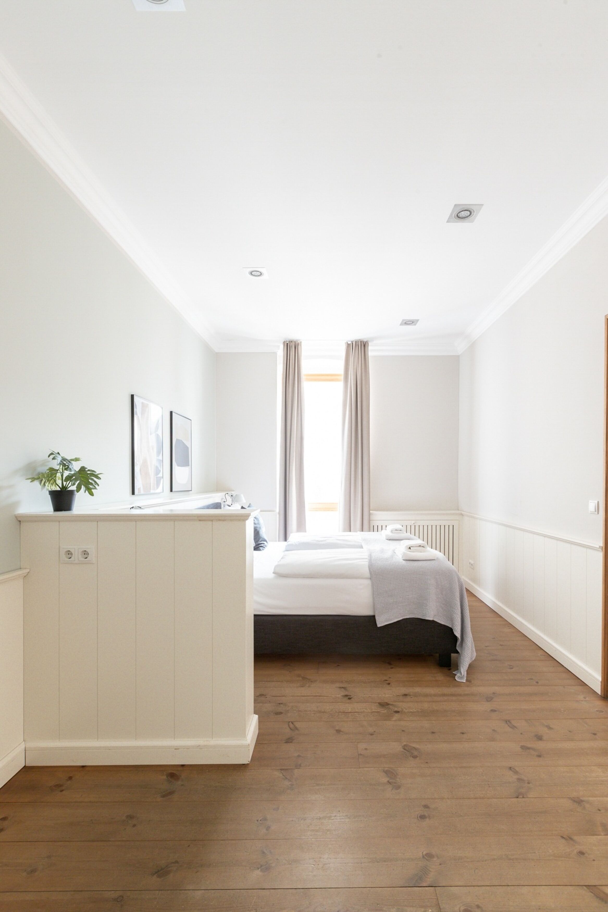 A bedroom with a bed, a bedside table, a radiator, a window with curtains, and a potted plant.