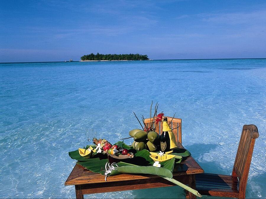 Een houten tafel met een tropisch fruitarrangement en een fles op een strand met helderblauw water en een eiland in de verte.