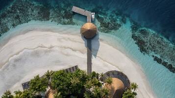 Vista aérea de un complejo turístico en la playa con un muelle de madera que conduce a una estructura de techo de paja, rodeado de palmeras y aguas turquesas cristalinas.