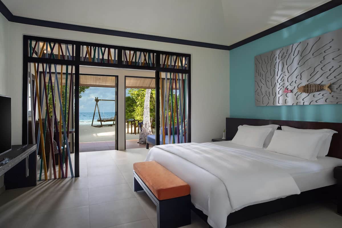 A modern hotel room with a large bed, a bench, a TV, and a view of the beach through a glass door.