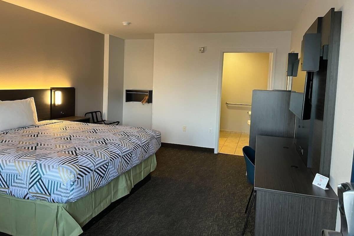 A hotel room with a bed, a desk, a chair, a TV, and a shower area.