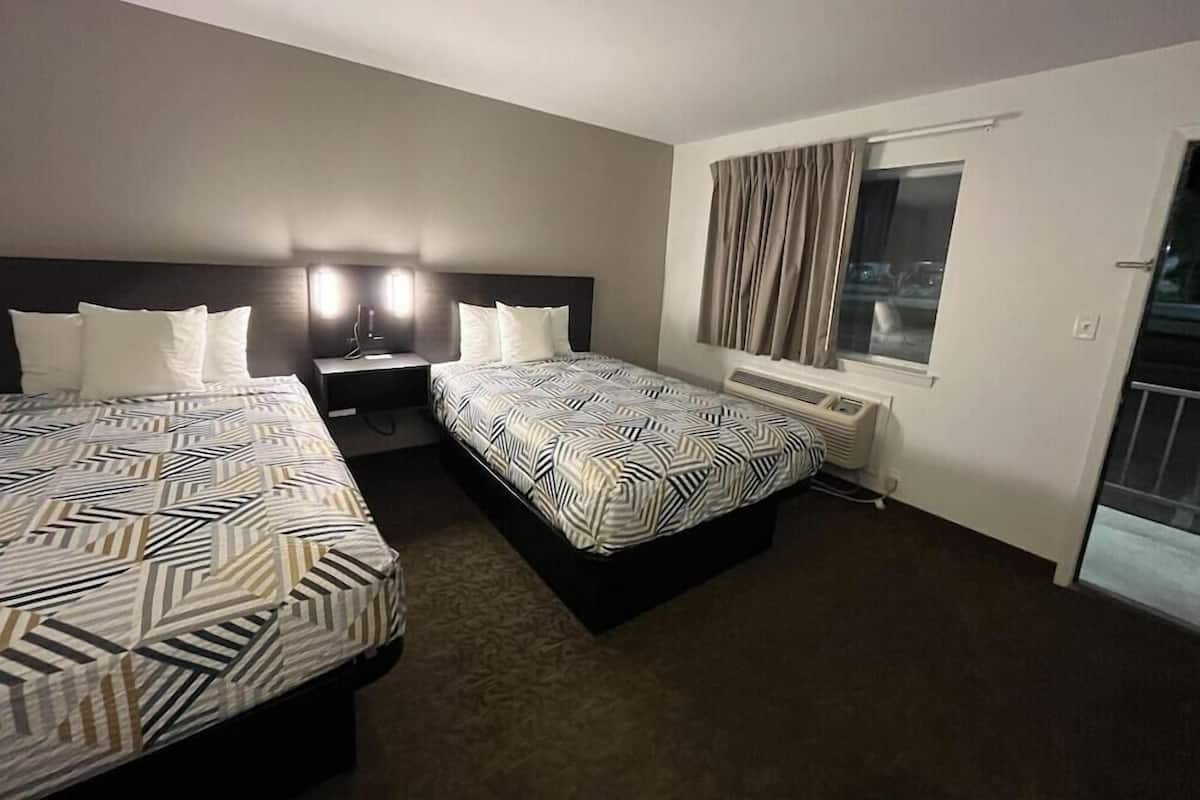 A hotel room with two beds, a nightstand, a window with curtains, and a door.