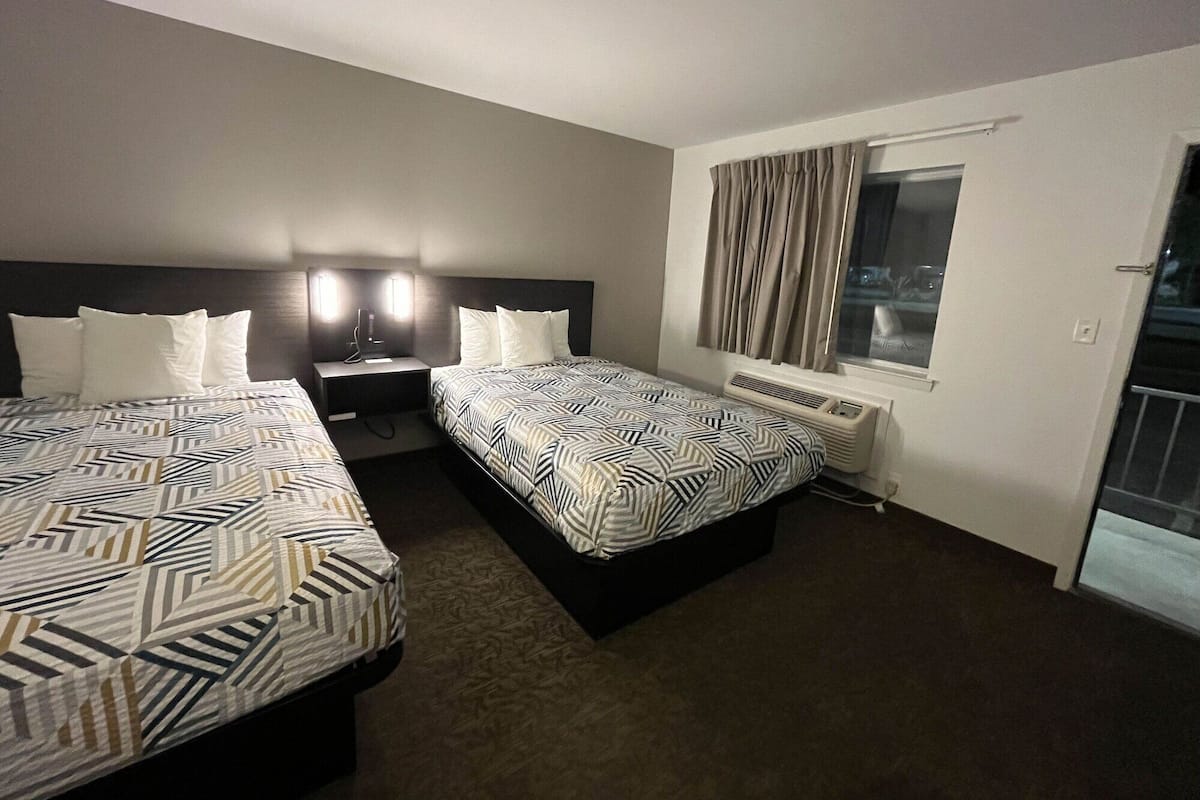 A hotel room with two beds, a nightstand, a heater, and a window with curtains.