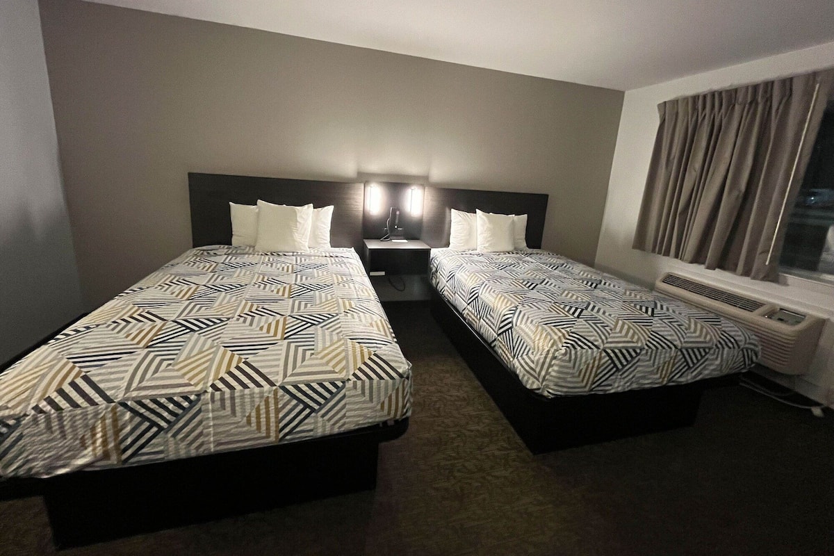 A hotel room with two beds, each with a patterned bedspread, a bedside table with a lamp, and a window with curtains.