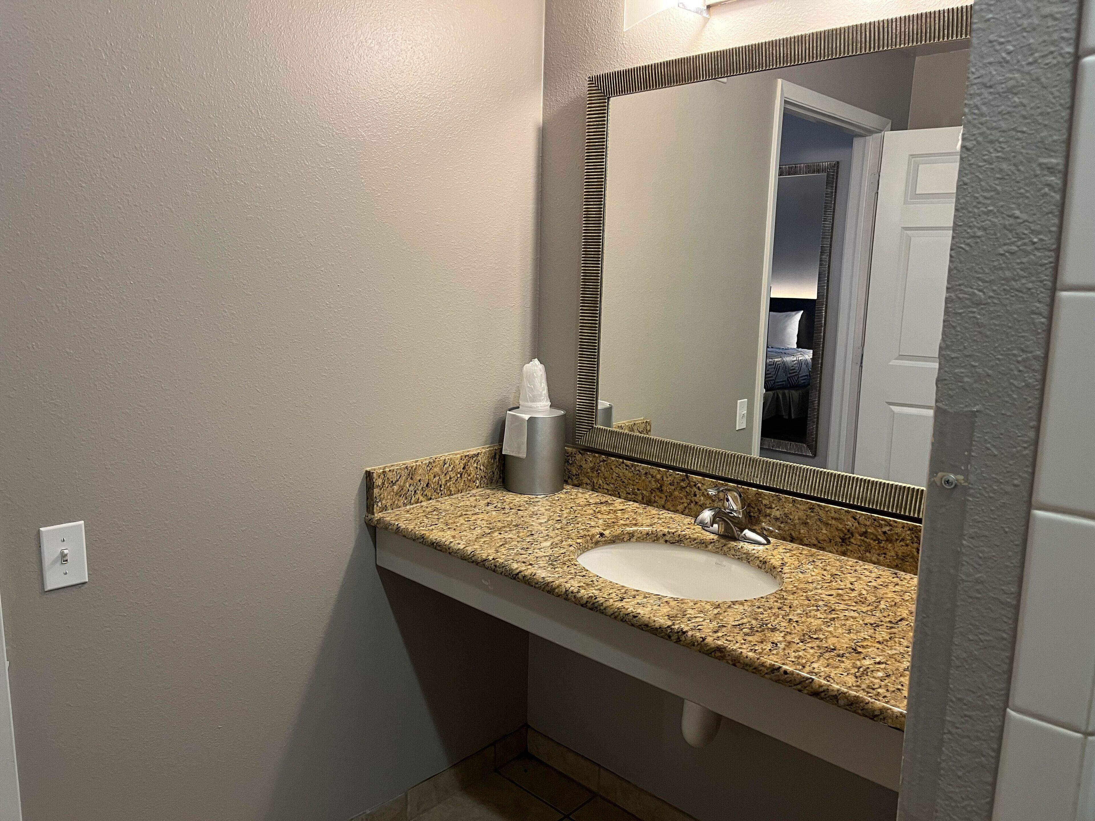 A bathroom with a granite countertop sink, a mirror, and a towel dispenser.