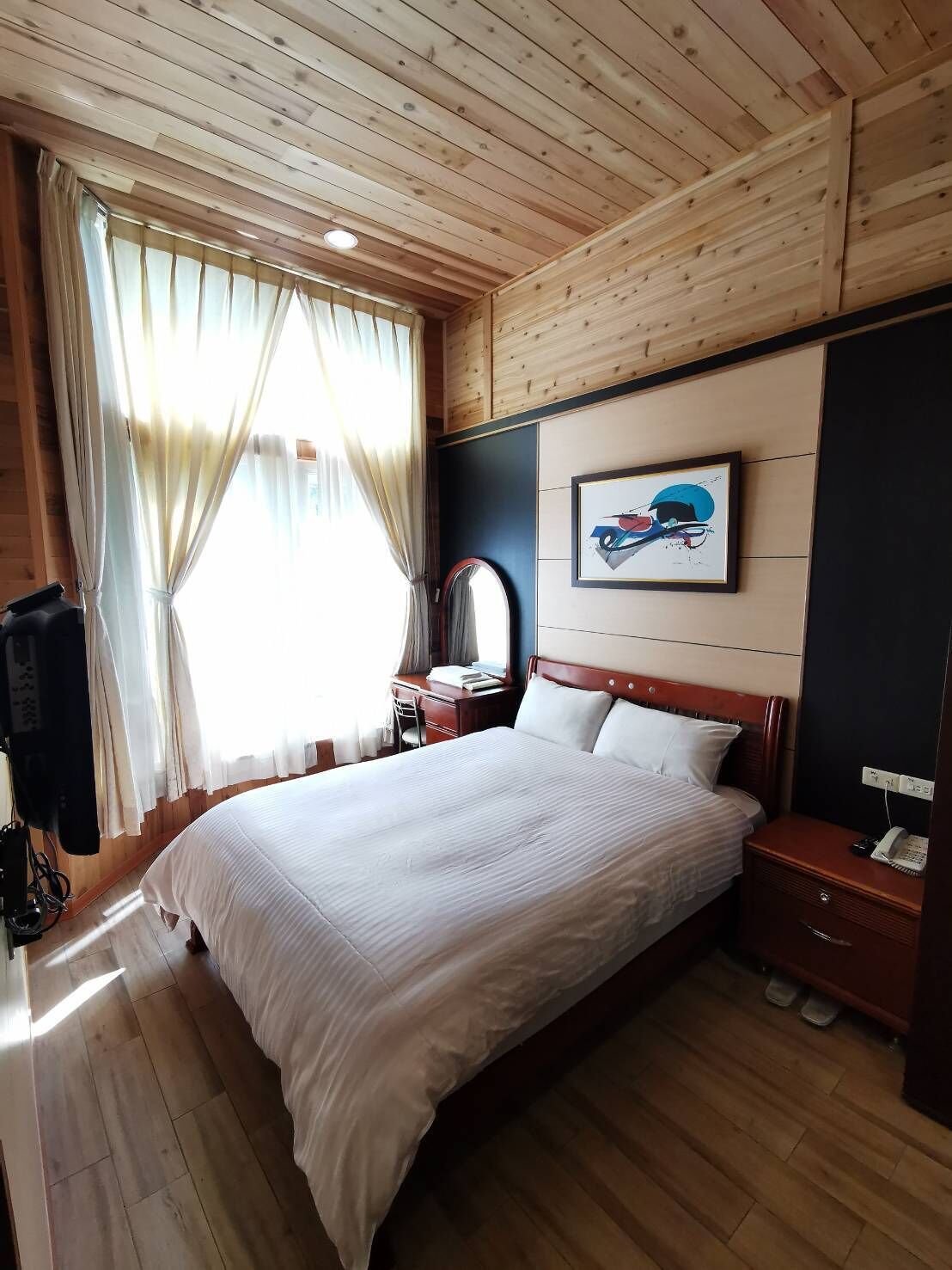 A wooden-paneled room with a large bed, a bedside table with a lamp, a mirror, and a painting on the wall.