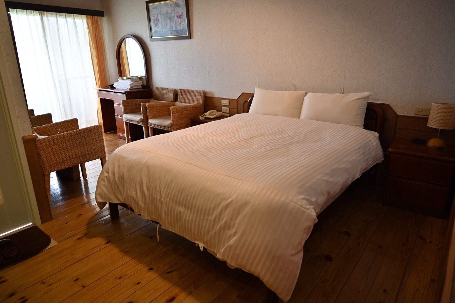 A bedroom with a large bed, a desk with a chair, a mirror, and a bedside table with a lamp.