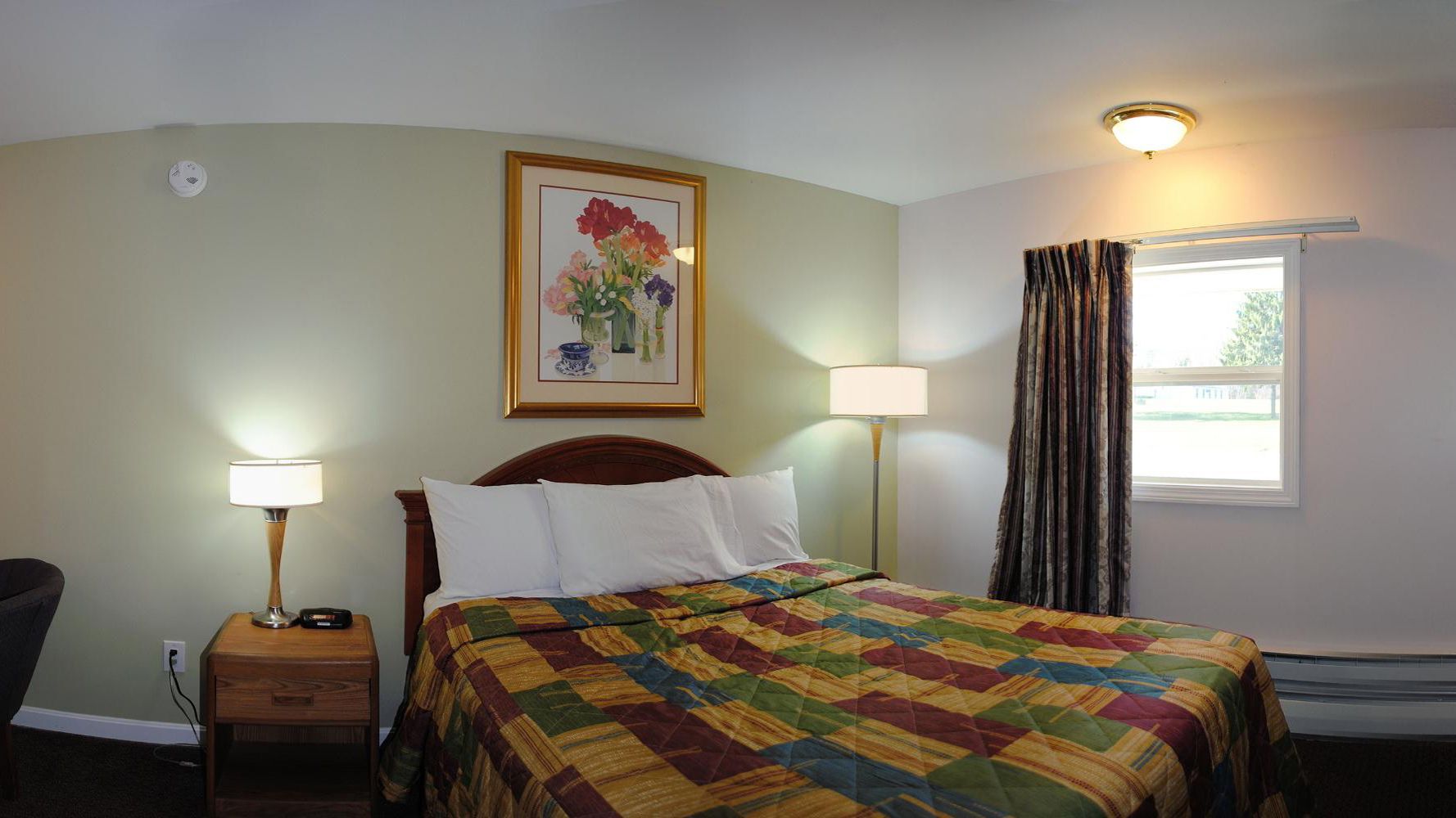 A hotel room with a bed, a nightstand with a lamp, a painting on the wall, and a window with curtains.