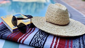 Escena junto a la piscina con un sombrero de paja, gafas de sol y un libro sobre una manta a rayas.