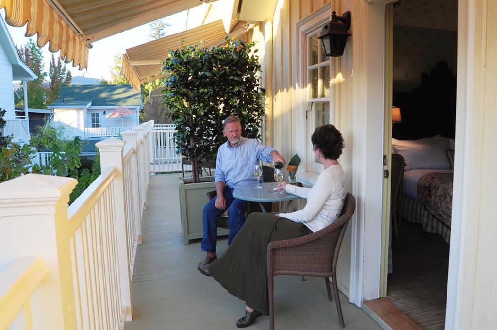 A covered porch with a dining table and chairs, a potted plant, and a view of a house in the background.