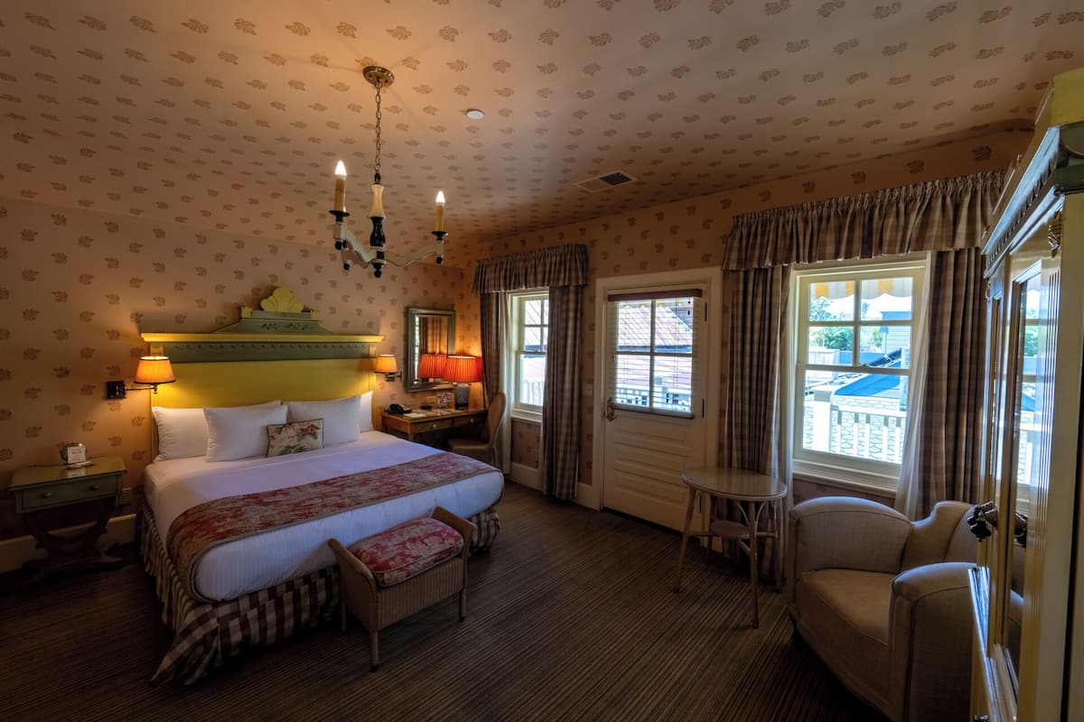 A hotel room with a large bed, a small table, a chair, a lamp, and a window with curtains.