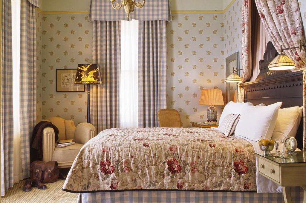 A hotel room with a large bed, a chair, a desk with a lamp, and a nightstand with a clock.
