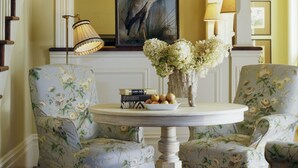 Una habitación con candelabro, sillones tapizados con flores, una mesa con un jarrón y un cuadro en la pared.