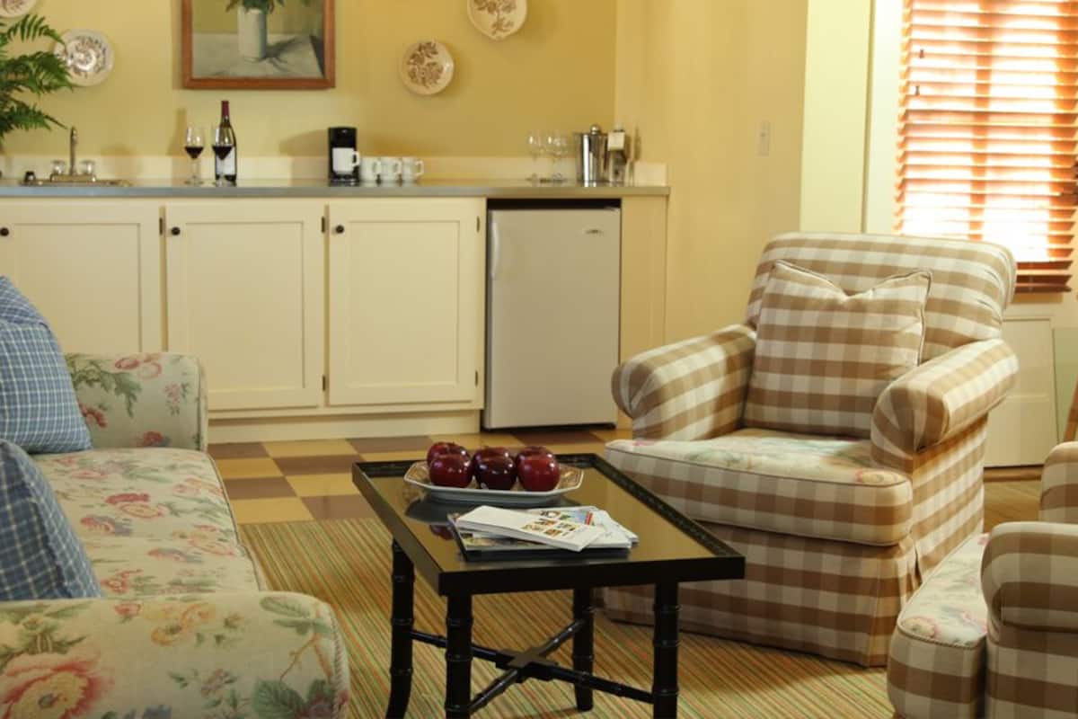 A cozy living room with floral-patterned sofas, a checkered armchair, a coffee table with a plate of apples, and a kitchen area with a refrigerator and a sink.