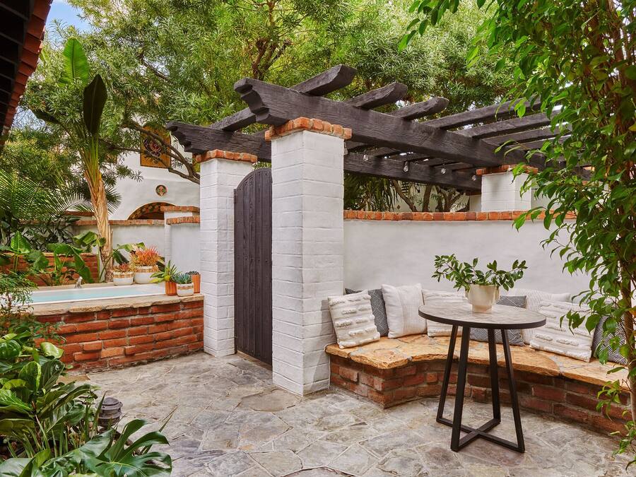 Eine Terrasse mit Steinboden, einer Holz-Pergola, einer Ziegelbank und einem kleinen Tisch mit einem Blumentopf.