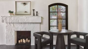 A room with a white fireplace, a round table with two chairs, and a large window with a view of greenery.