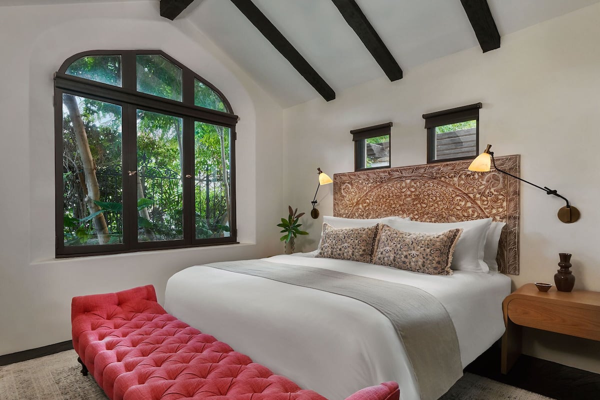 A bedroom with a large window, a bed with patterned pillows, a red bench, and a nightstand with a lamp.