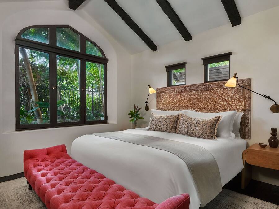 Un dormitorio con un ventanal grande, una cama con almohadones estampados, un banco rojo y una mesita de noche con una lámpara.