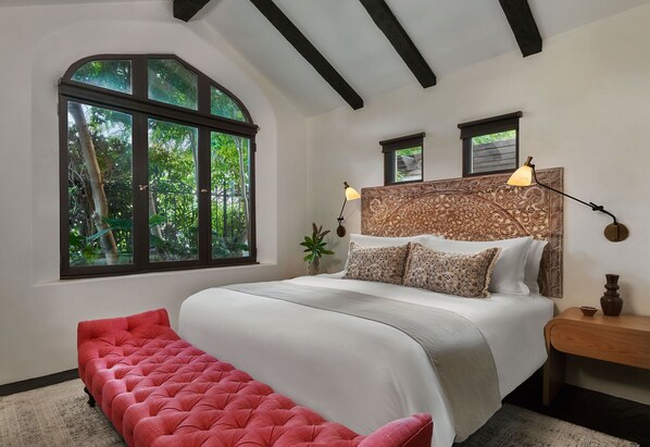A bedroom with a large window, a bed with patterned pillows, a red bench, and a nightstand with a lamp.