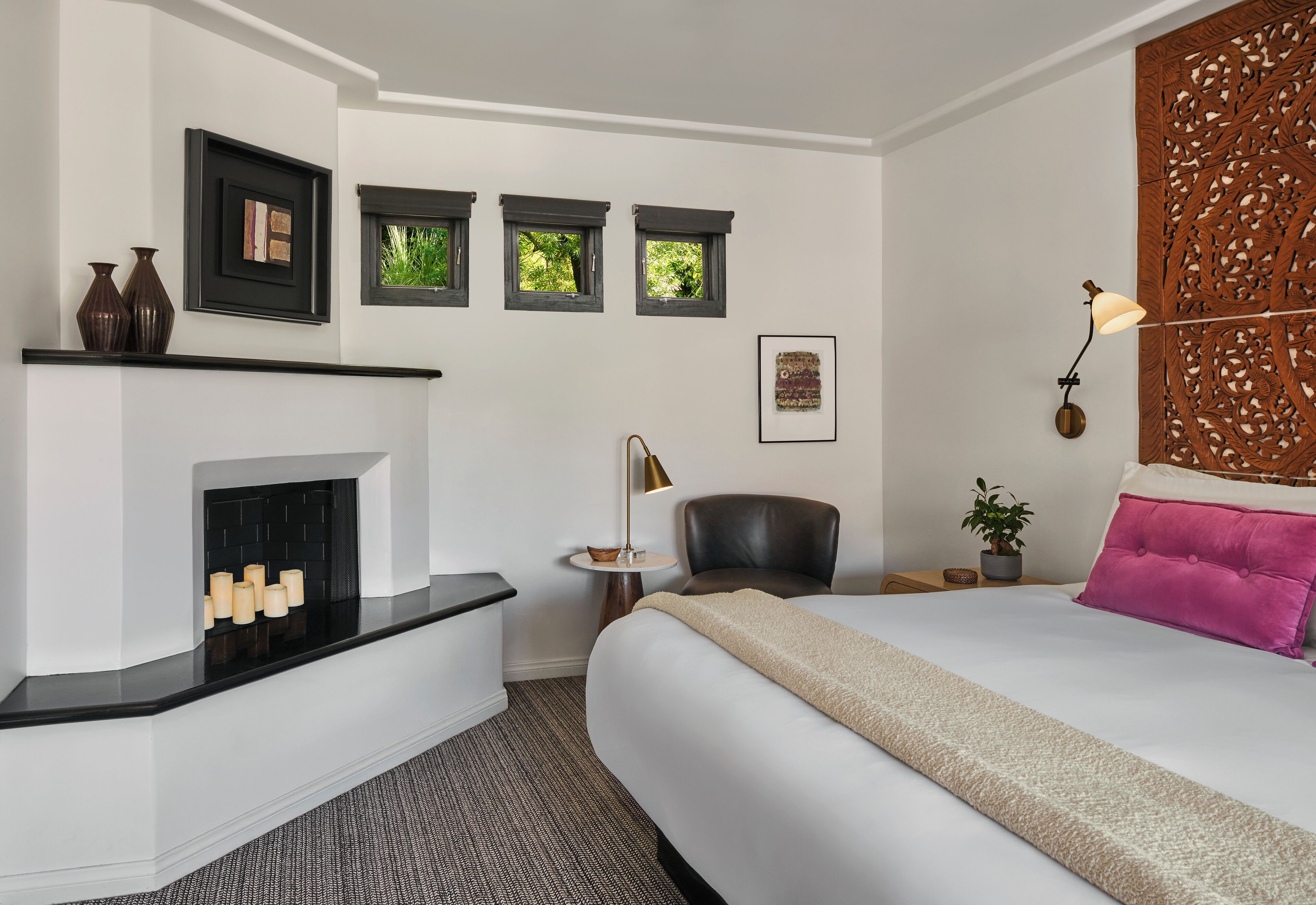 A hotel room with a fireplace, a bed with a beige blanket, a chair, a small table with a lamp, and a decorative wall panel.